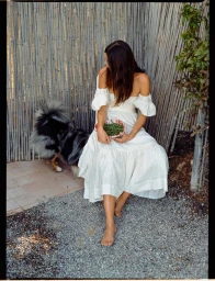 Woman in white off-shoulder dress holding a bowl seated next to a fluffy dog by a bamboo fence.