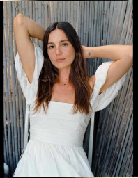 Portrait of a woman with dark hair in a white dress posing against a vertical bamboo fence.