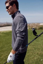Man wearing sunglasses and grey pullover holding a golf club on a green course with a golf bag in the background.