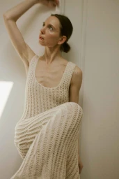 Woman in cream knit dress posing with raised arm against a pale paneled wall