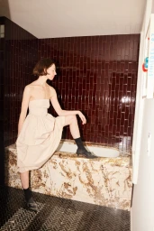 Woman in cream strapless dress with dark socks and heels posing with one leg on a stone bathtub lined with dark tile.