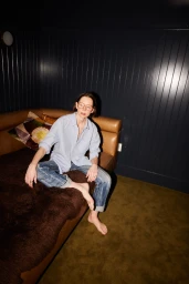 Woman wearing glasses, light blue button-down shirt and denim jeans seated on a brown leather sofa in a dark paneled room