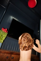 A young child with red hair reaching up toward a red balloon inside a dark-paneled room with a vase of tulips.