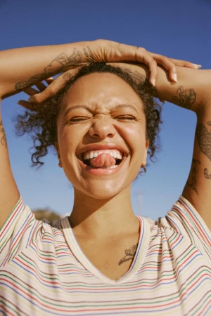 Portrait of a woman with curly hair sticking her tongue out, wearing a striped t-shirt against a clear blue sky background.