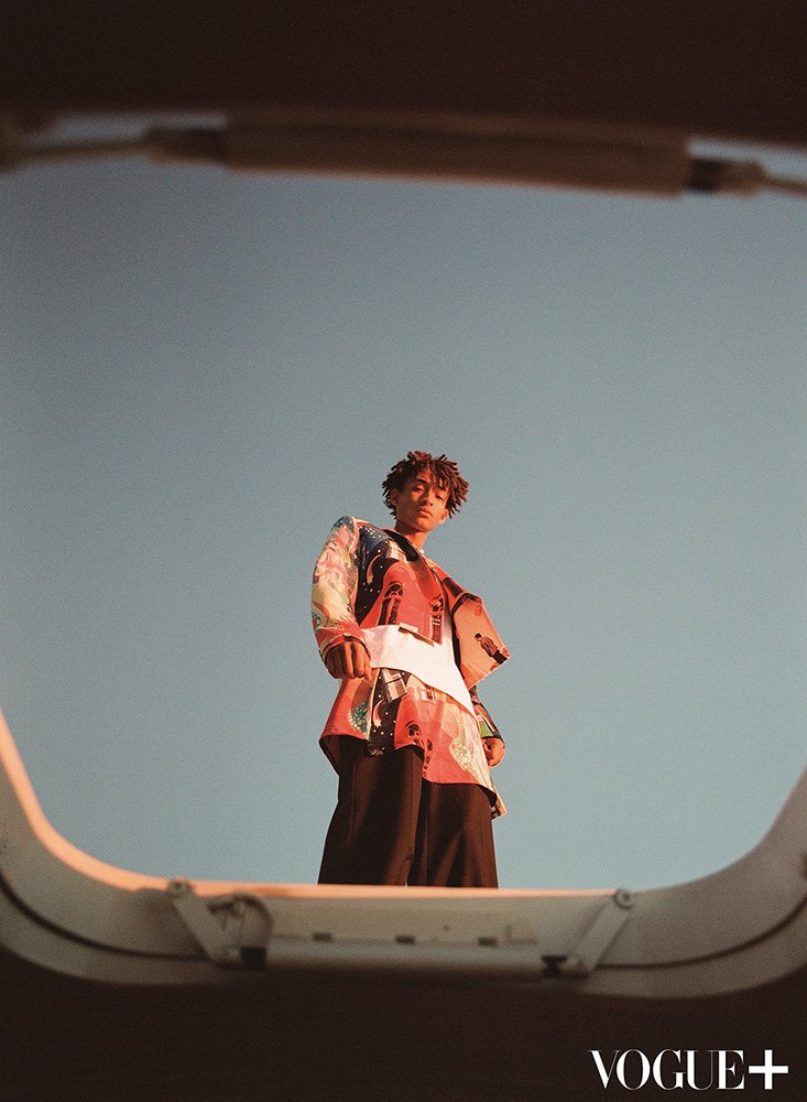 A stylish person with medium brown skin and curly hair, wearing a printed top and pants, standing on a rooftop against a blue sky.