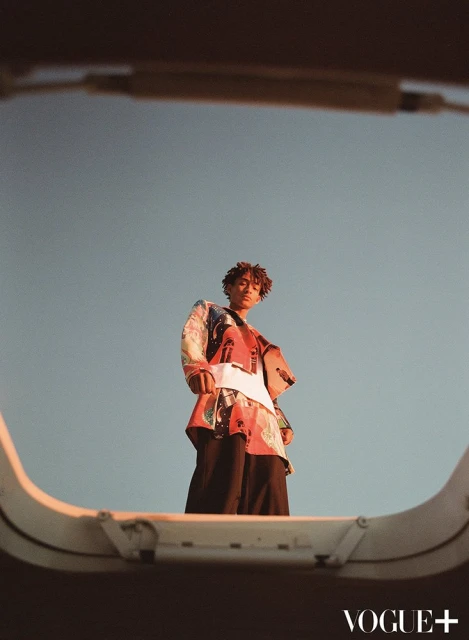 A stylish person with medium brown skin and curly hair, wearing a printed top and pants, standing on a rooftop against a blue sky.