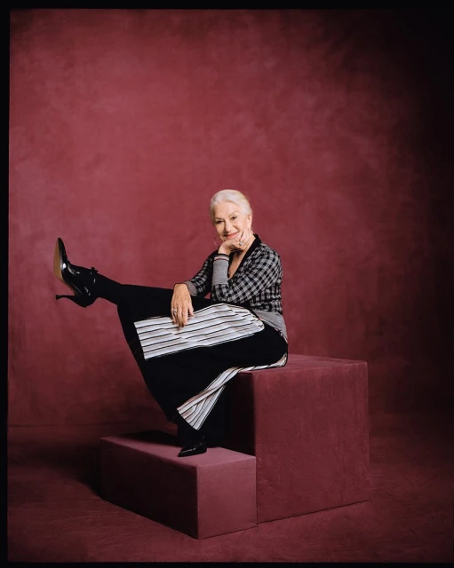 Mature woman poses elegantly on mauve platforms, wearing a checkered top, flowing skirt, and shiny heeled boots against a matching backdrop.