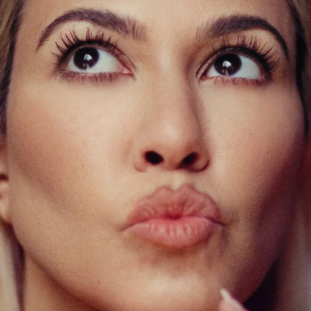 Close-up portrait of a young adult woman with brown eyes, long eyelashes, arched eyebrows, and pursed lips against a soft-focus background.