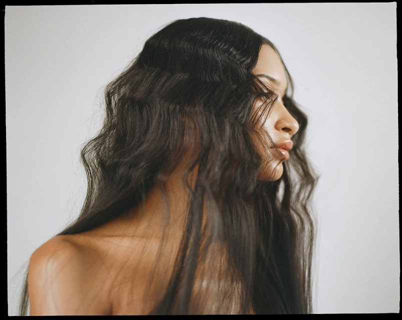 Profile portrait of a bare-shouldered woman with long dark wavy hair in a light studio. Beauty and fashion editorial.