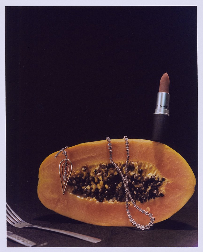 Still life of a halved papaya with a necklace, lipstick, and silverware against a dark background, showcasing beauty and food fusion.