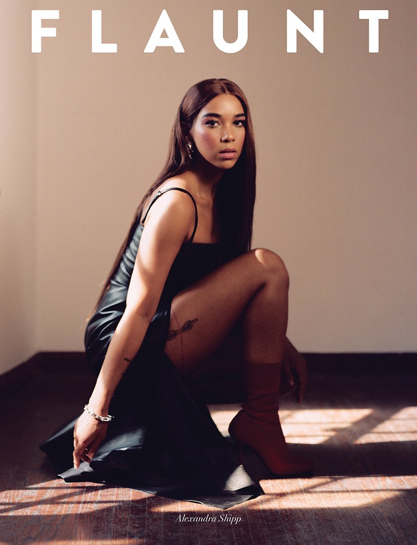 A woman in a black dress and brown boots poses in a studio setting. The image includes the 'Flaunt' magazine logo and the name Alexandra Shipp.