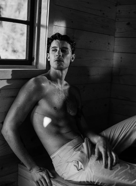 Black and white portrait of a shirtless adult male in light-colored pants relaxing against wood wall and window in indoor setting.