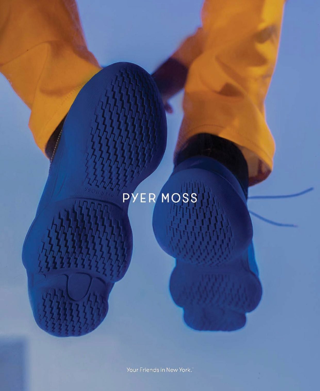 Angled low shot of a person wearing yellow pants and blue sneakers from below, showing the soles. "PYER MOSS" brand is visible.