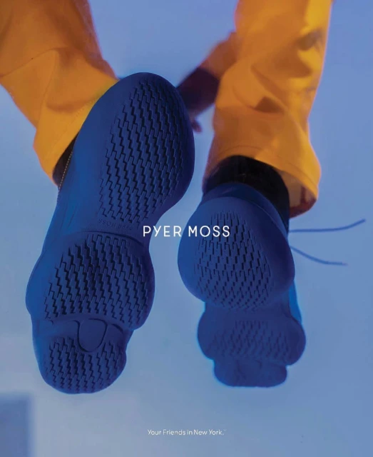 Angled low shot of a person wearing yellow pants and blue sneakers from below, showing the soles. "PYER MOSS" brand is visible.