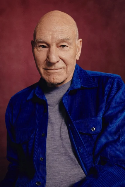 Portrait of Patrick Stewart. He has a bald head, and he is wearing a grey top and a blue corduroy jacket. Burgundy background.