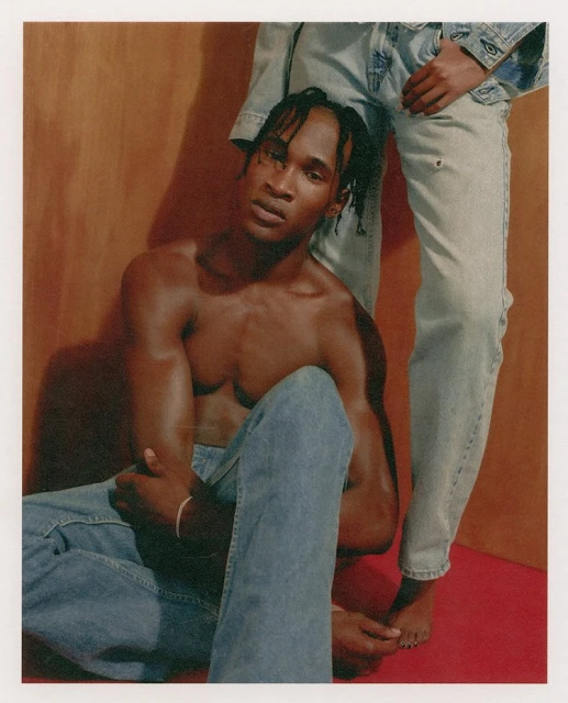 Two people pose in a studio with earth-tone background. One shirtless man sits in denim jeans, while another wears a denim outfit.
