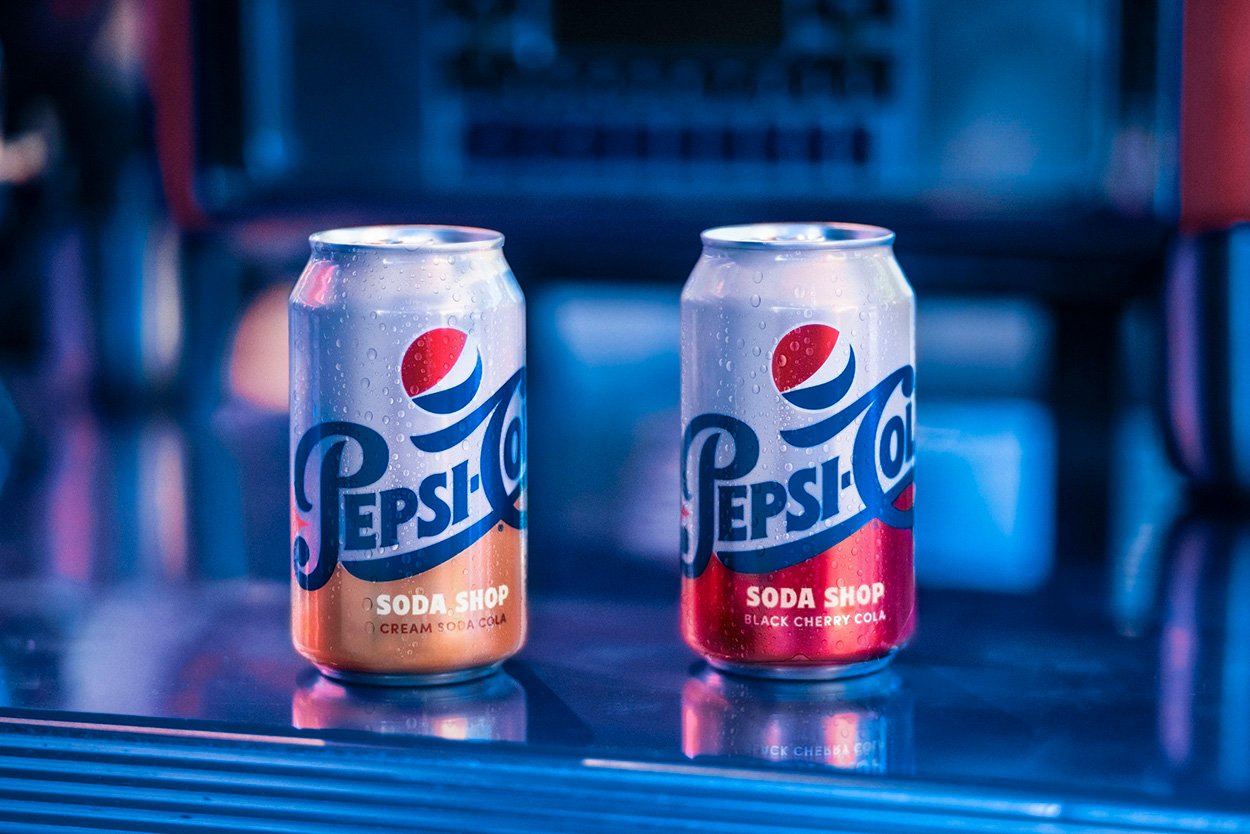 Two Pepsi-Cola Soda Shop cans sitting on a reflective blue surface under moody neon lighting.
