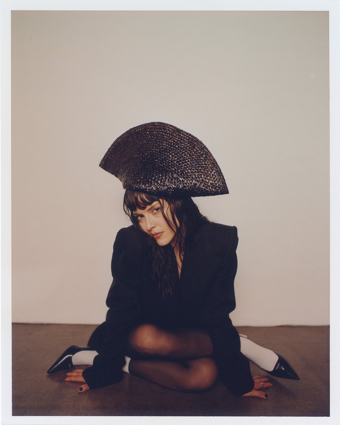 Stylish portrait of a female model in a black blazer and dark textured hat, sitting on the floor in front of a white wall.