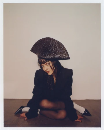 A woman in a black blazer and structured fan-shaped hat sits cross-legged against a neutral wall in a polaroid portrait.
