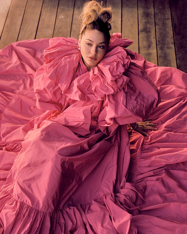 Woman in a voluminous pink dress poses on a wooden surface, creating a striking fashion portrait with ruffles and texture.