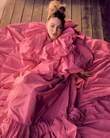 Woman with an updo hairstyle wearing a voluminous ruffled pink gown while seated on a wooden plank floor.