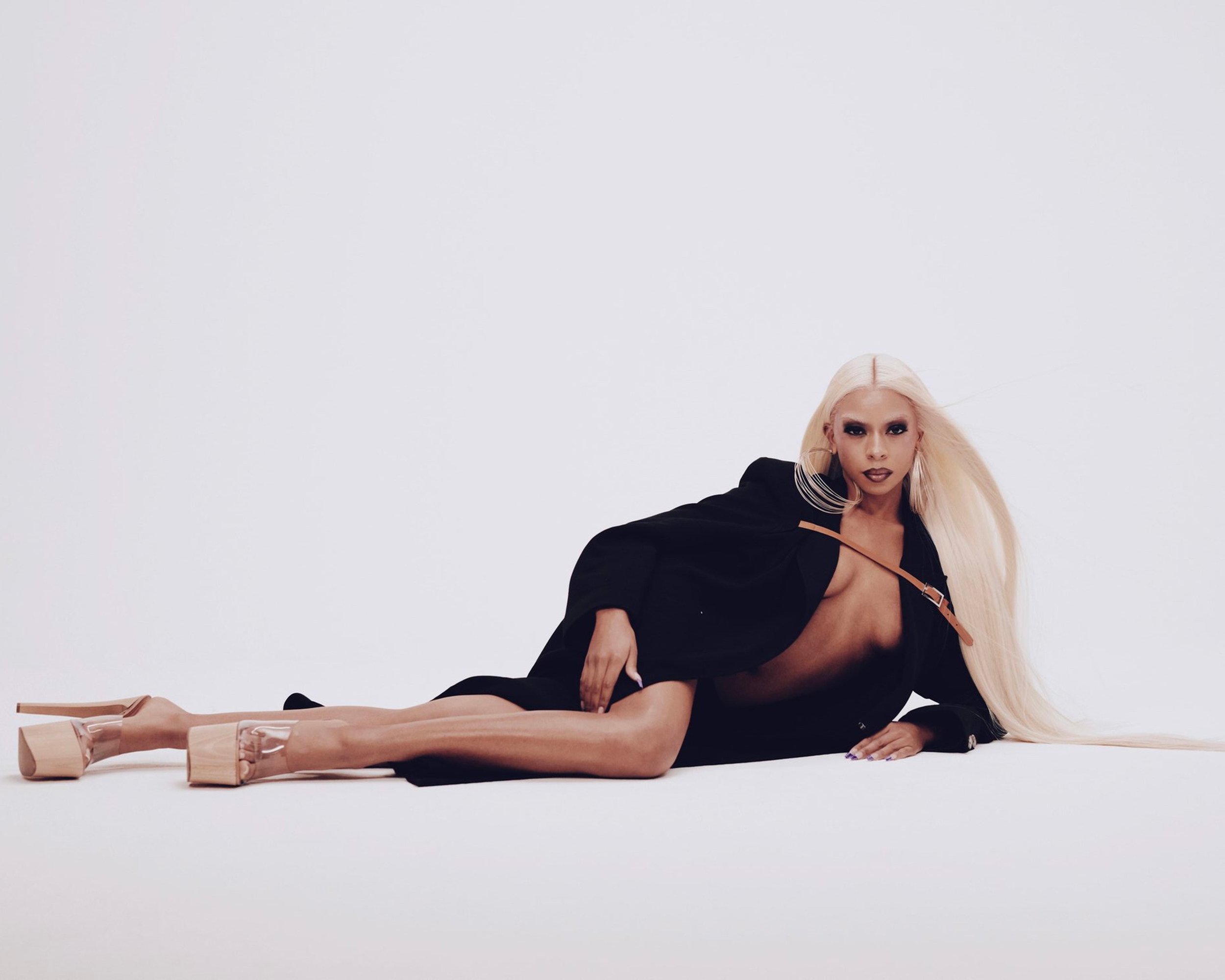 Model with long blonde hair wearing a black coat and platform heels lying on a white seamless background
