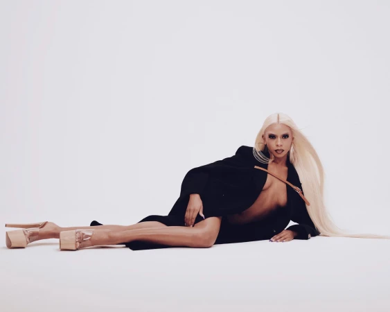 Model with long blonde hair wearing a black coat and platform heels lying on a white seamless background