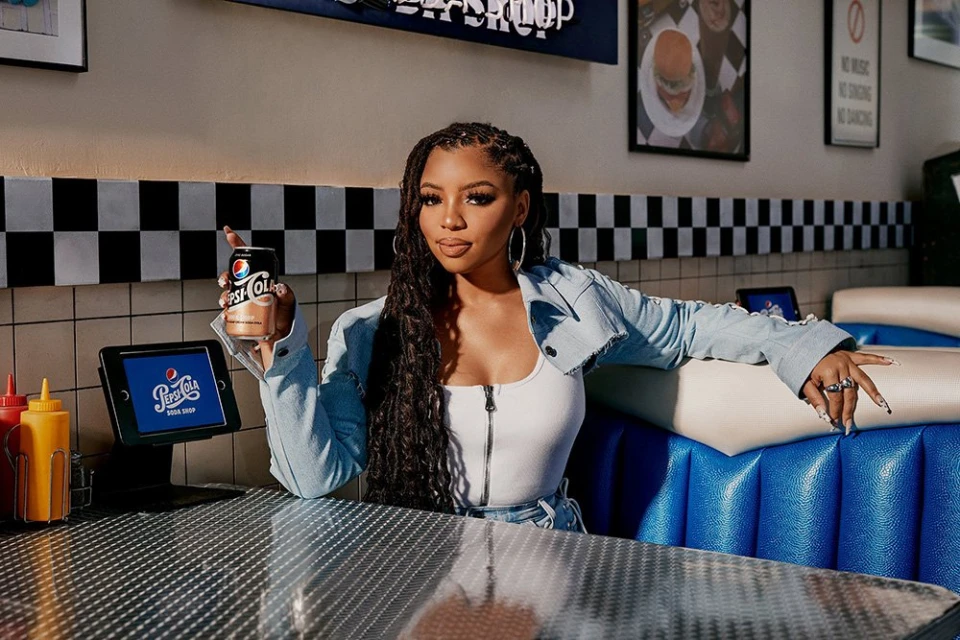 A young woman in a diner booth holds a Pepsi can, with a checkerboard wall and soda-shop details creating a vintage atmosphere.