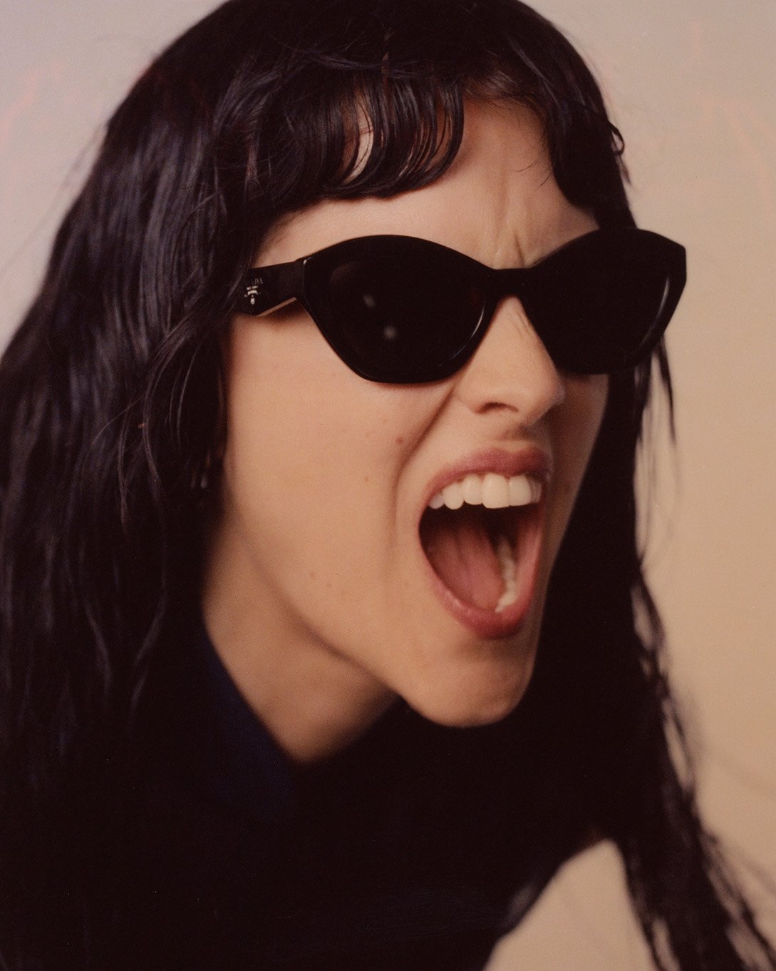 Close-up of an adult woman with dark hair and sunglasses, mouth open, yelling, wearing a dark shirt, with a neutral background.
