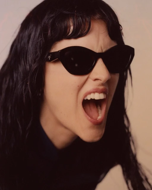 Close-up of an adult woman with dark hair and sunglasses, mouth open, yelling, wearing a dark shirt, with a neutral background.