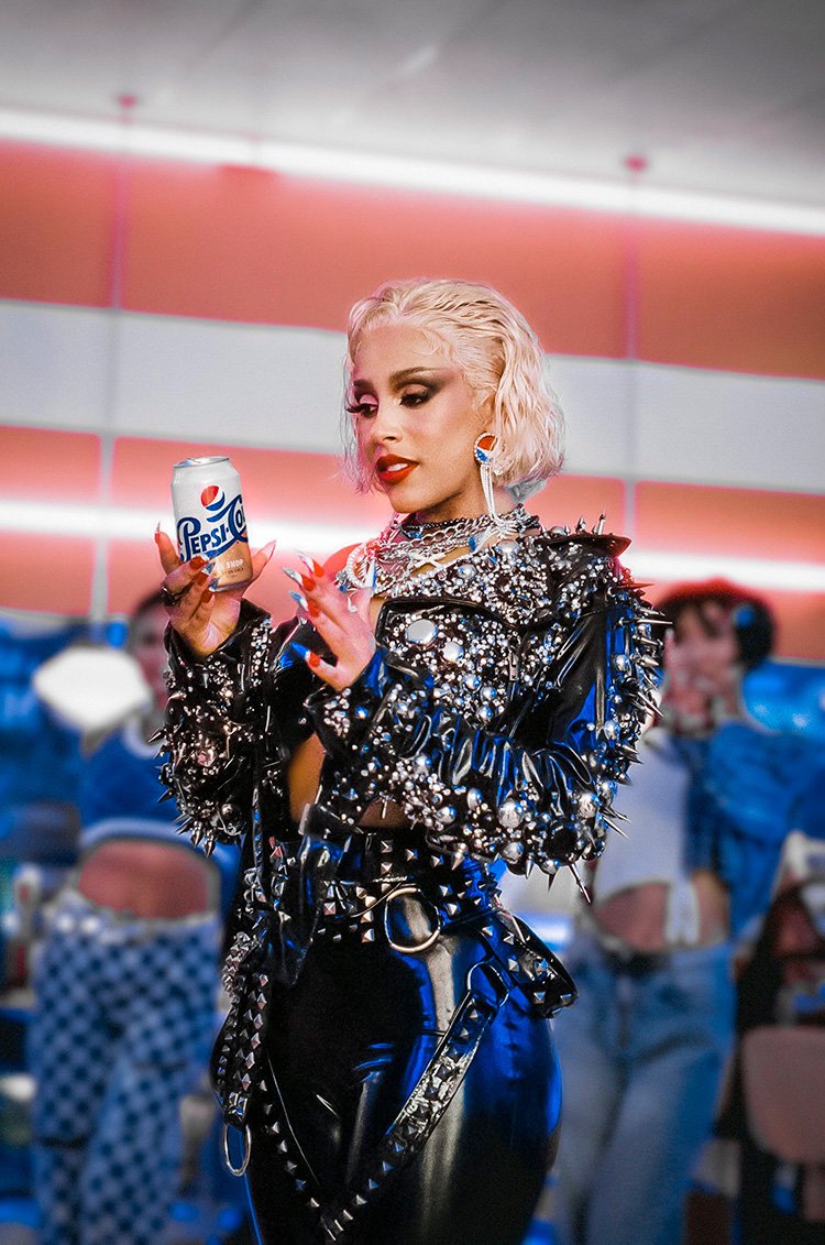 Adult female with platinum blonde hair holds a Pepsi-Co soda can, wearing an embellished punk-style jacket, in a studio environment.