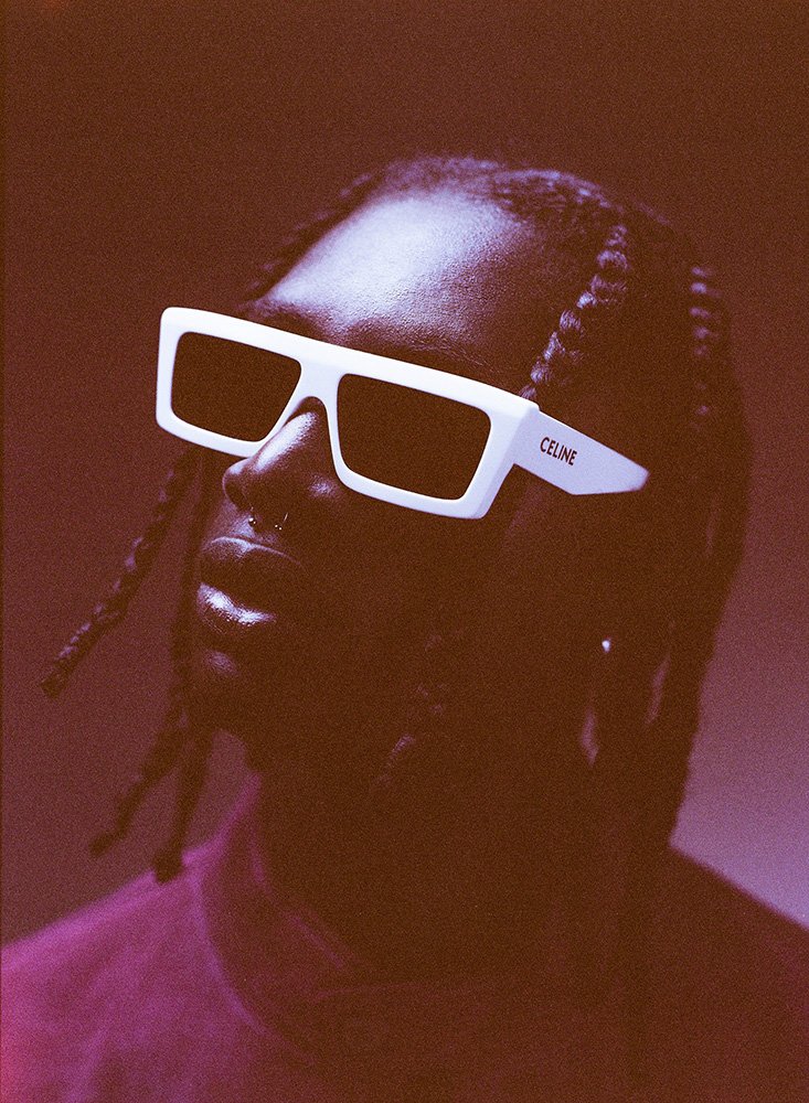 Close-up portrait of a black man wearing thick white frame sunglasses with braided hair against a reddish monochrome background
