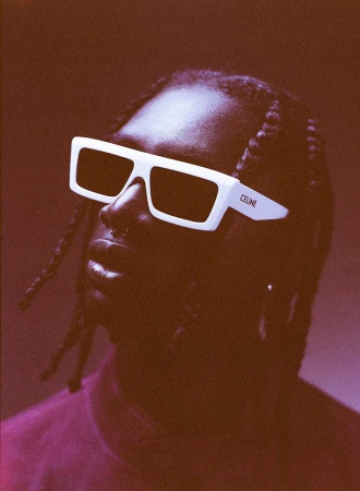 Close-up portrait of a black man wearing thick white frame sunglasses with braided hair against a reddish monochrome background