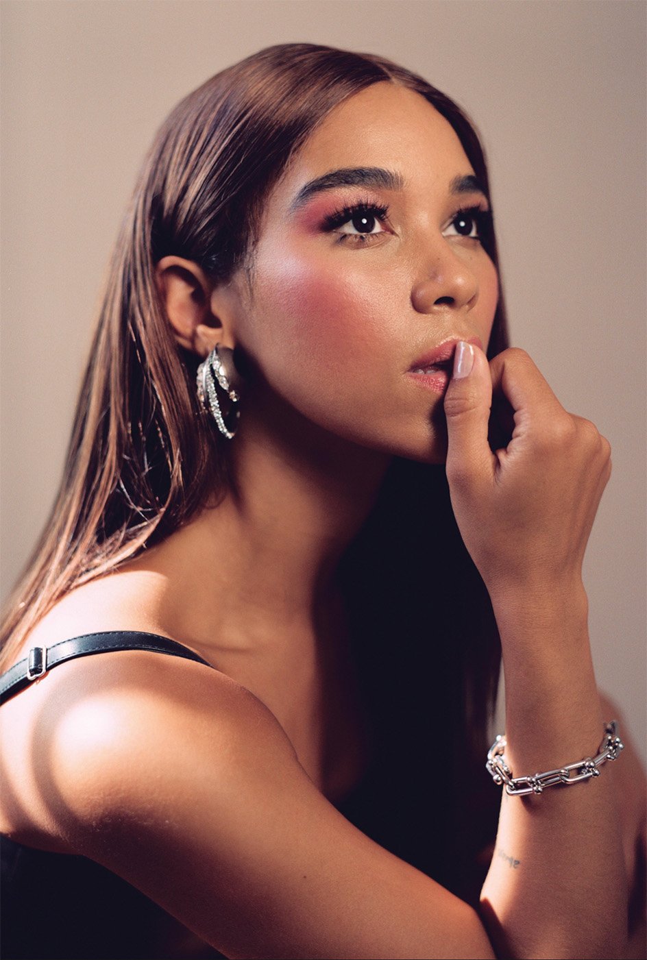 Close-up portrait of a woman with long brown hair, wearing sparkly hoop earrings and a bracelet, against a neutral background.