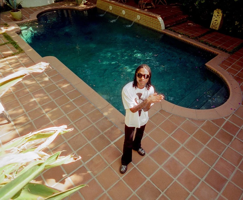 Eye-level shot of a man with sunglasses and braids standing by a swimming pool, wearing a white t-shirt and black pants outdoors.
