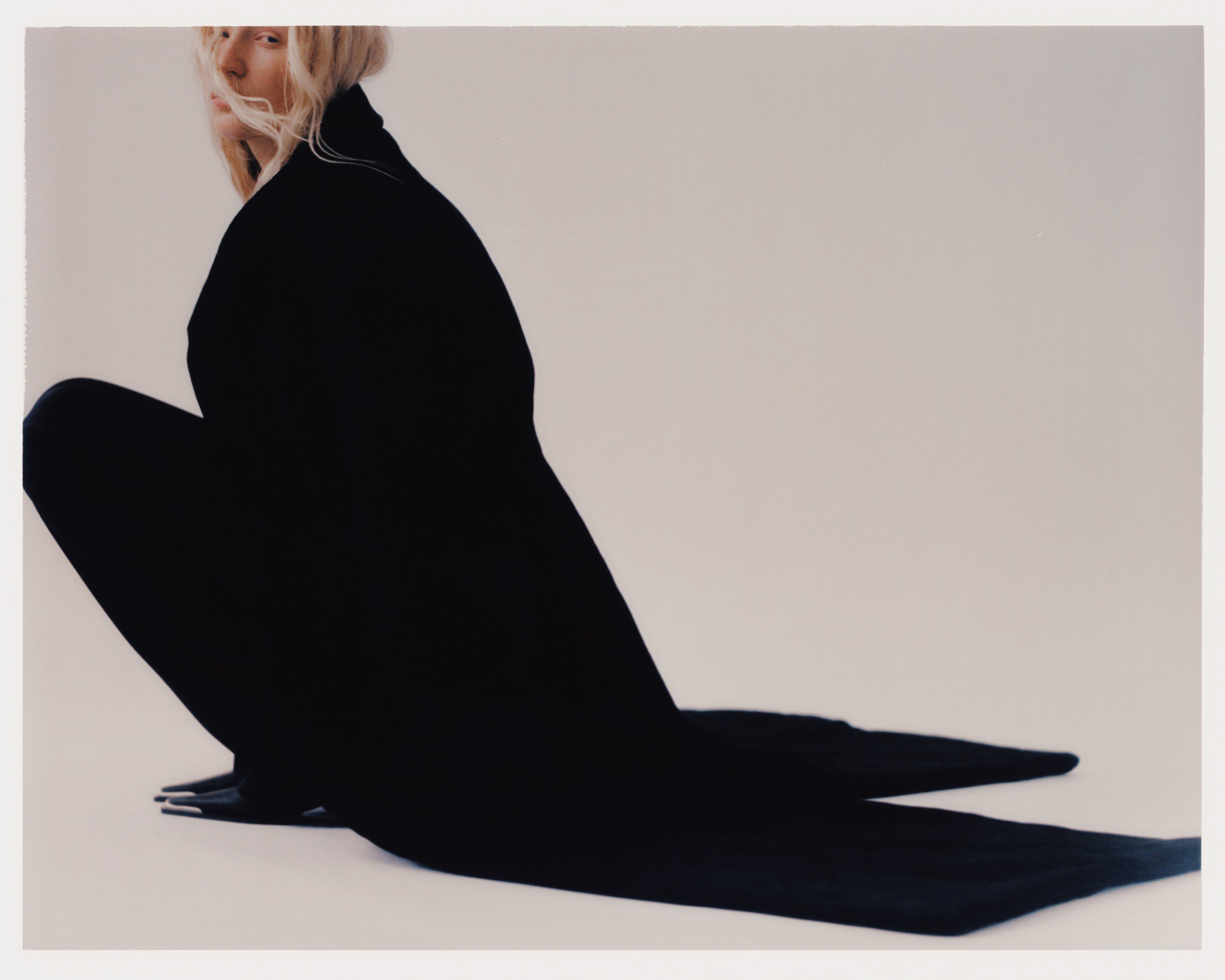 Fashion shot of an adult woman in a dark, full-body garment, crouching against a plain white backdrop in a photo studio.