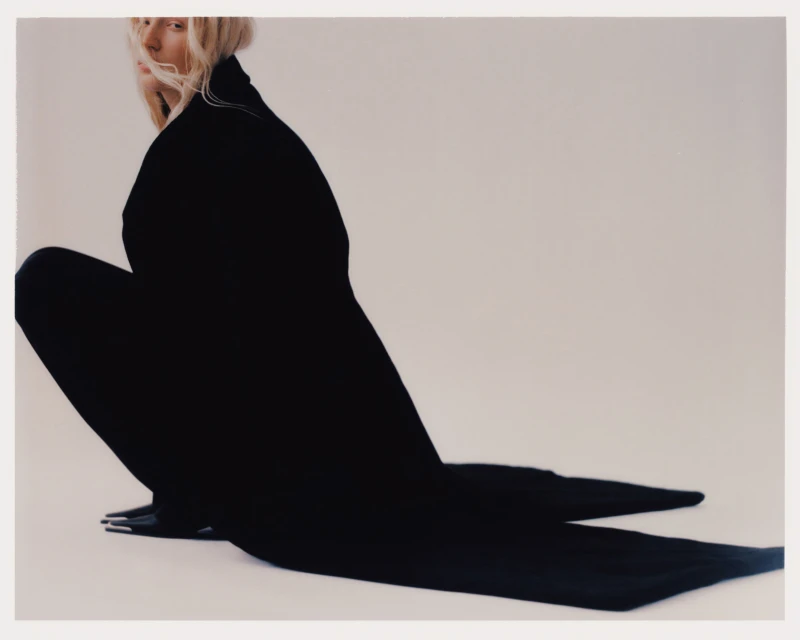 Fashion shot of an adult woman in a dark, full-body garment, crouching against a plain white backdrop in a photo studio.