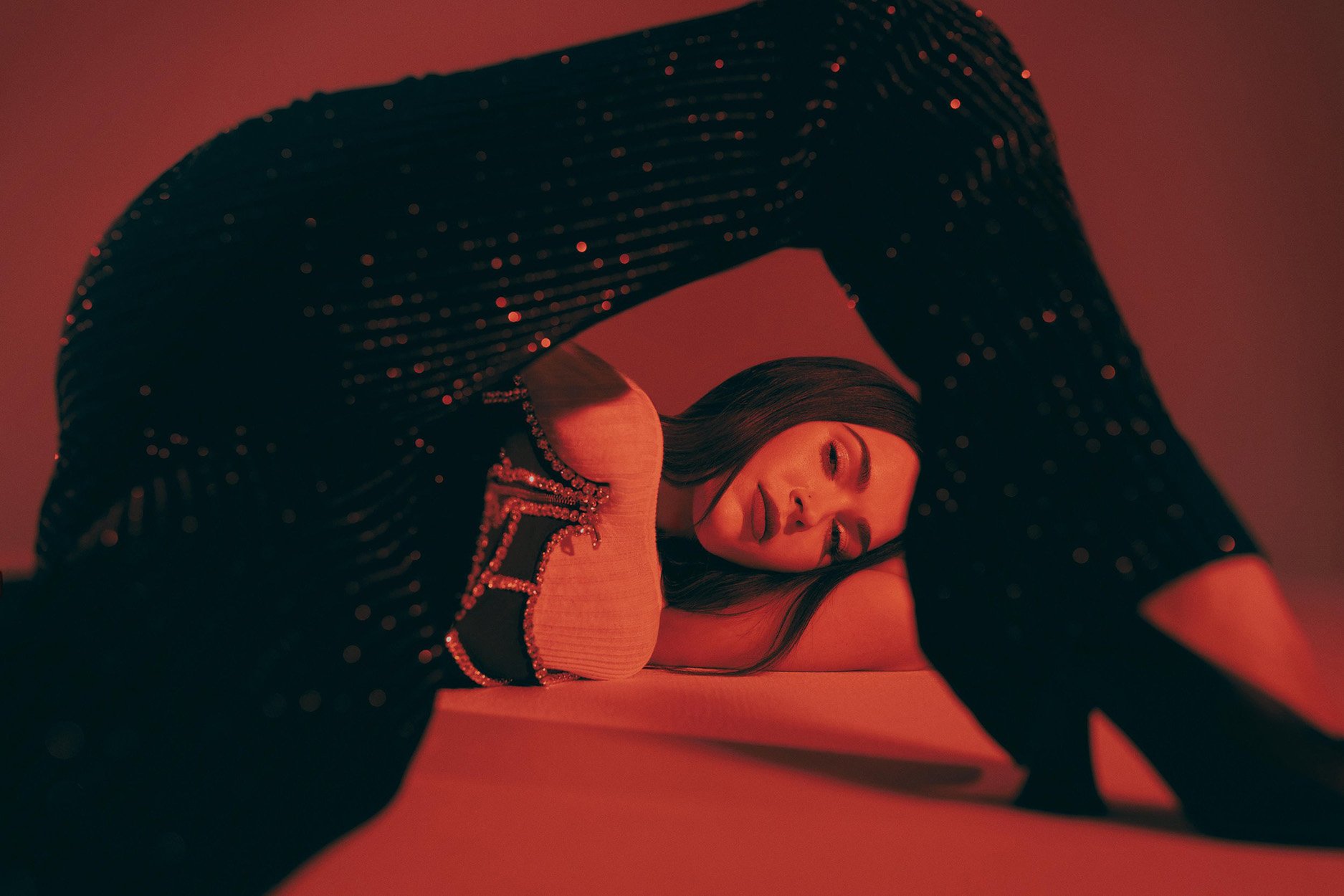 Woman in glittering jumpsuit poses with arched back on a red backdrop, showcasing a glamorous and dramatic fashion editorial.