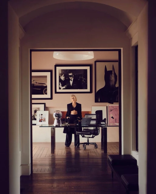 Woman in a black suit stands at a desk with laptop in a room with framed artwork, viewed through a hallway with white columns.
