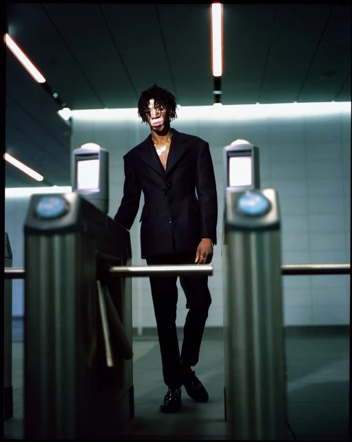 A young adult with dreadlocks in a black suit walks through a metal turnstile indoors under fluorescent lighting in a subway-like setting.