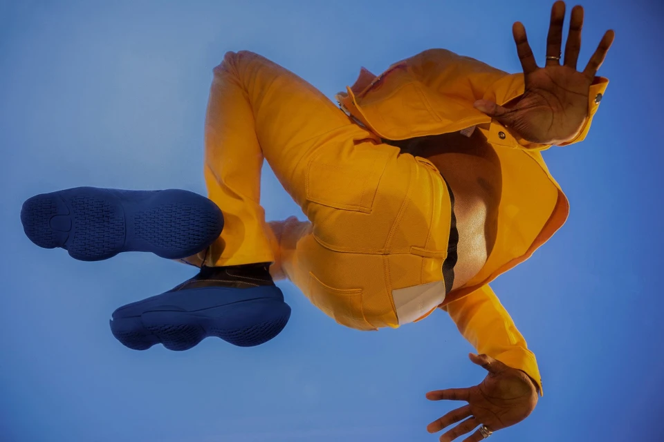 Low-angle shot of adult male wearing yellow outfit and blue shoes against a blue sky, arms raised in a dynamic pose.