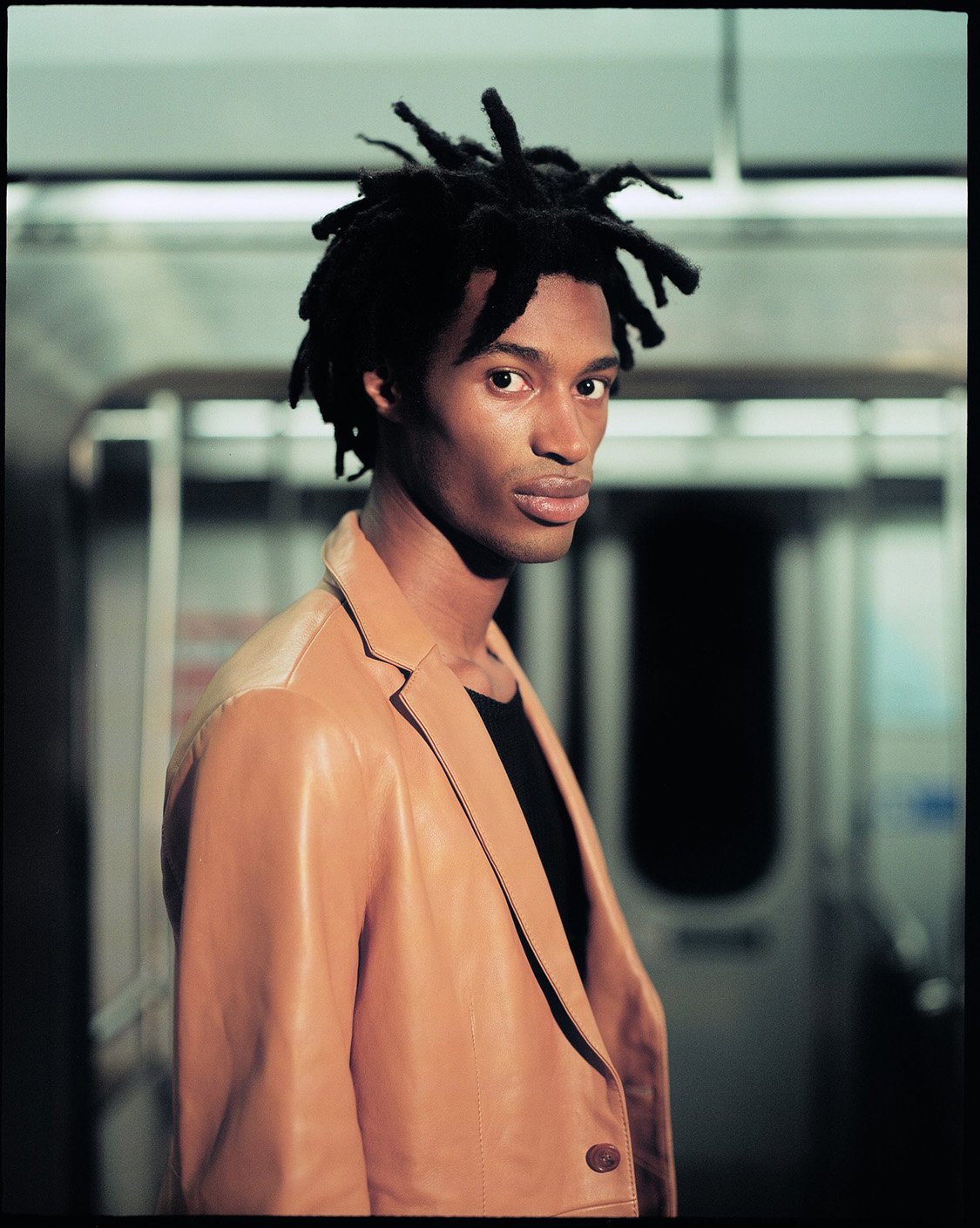 Portrait of a Black man with dreadlocks wearing a tan blazer inside of a subway car with gray walls, looking slightly off-center.