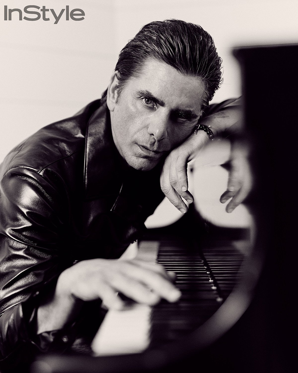 Black and white portrait of a man in a leather jacket leaning on a piano, InStyle magazine logo visible in the top left corner.