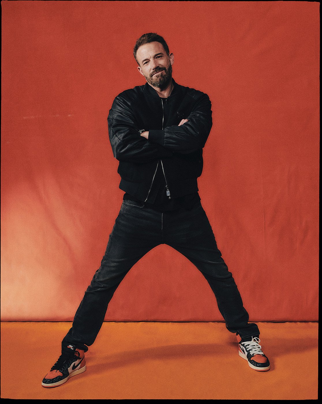 Adult man with a beard poses in a black jacket and jeans against an orange background, wearing black and orange sneakers. Arms are crossed.