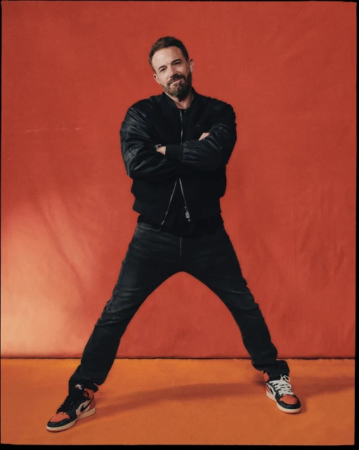 Adult man with a beard poses in a black jacket and jeans against an orange background, wearing black and orange sneakers. Arms are crossed.