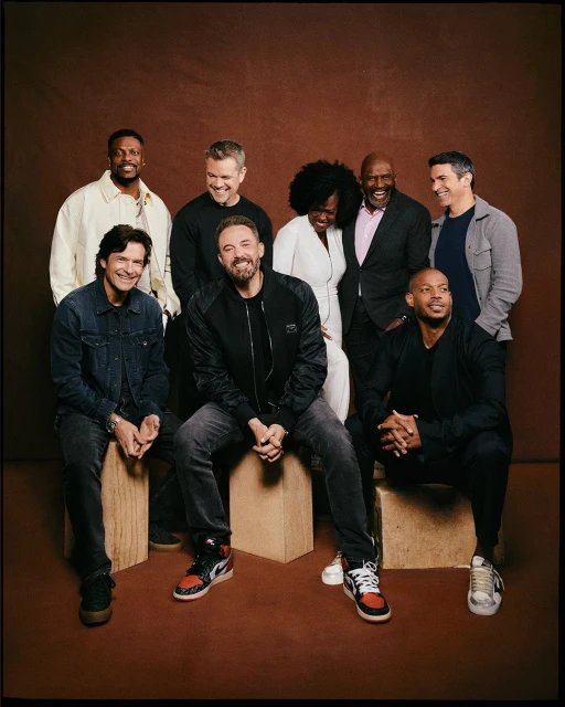 Group portrait of seven actors, including men and women, posing against a brown backdrop in a photo studio setting with soft lighting.