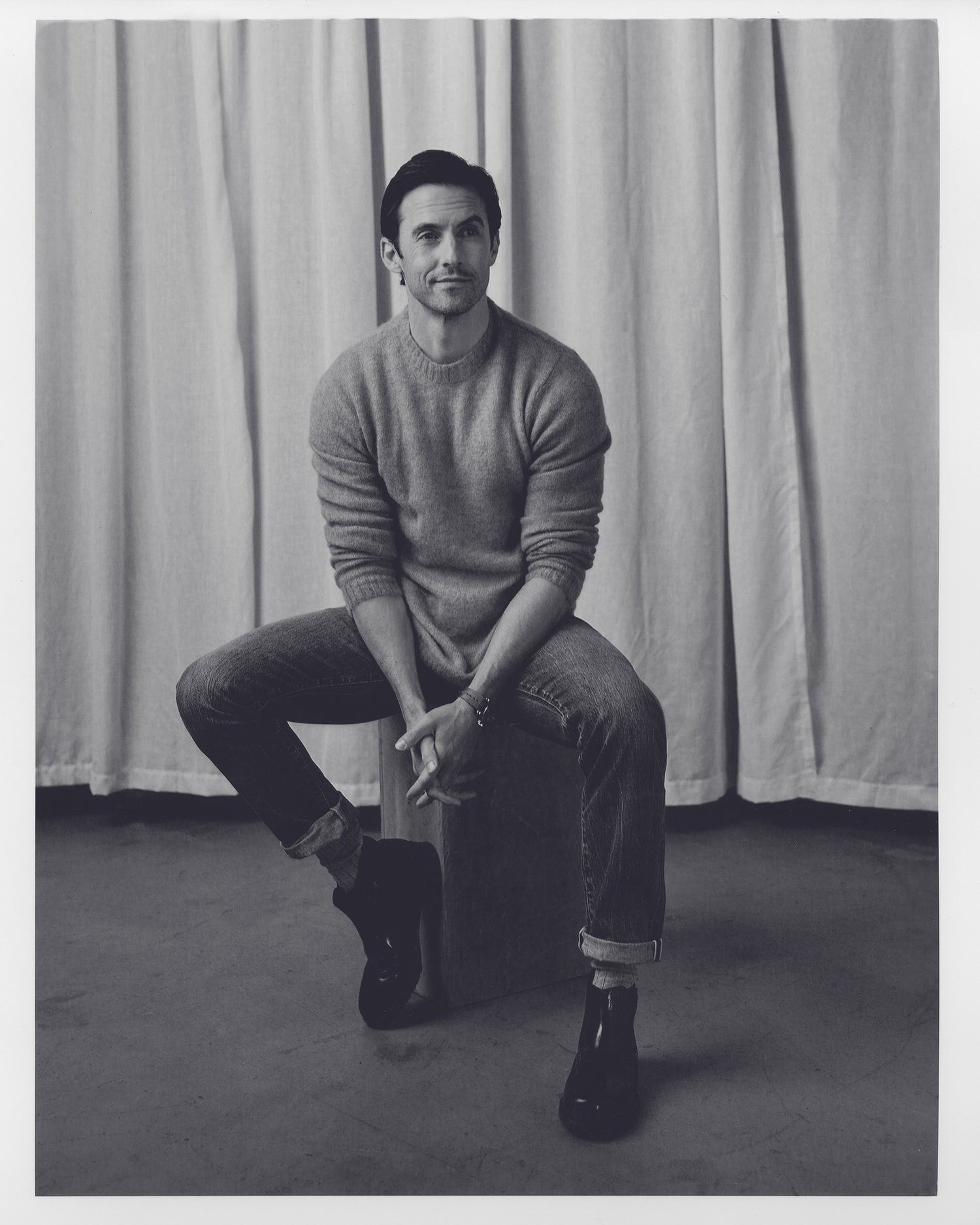 Portrait of a man in a crewneck sweater and cuffed jeans seated against a draped curtain background by Alex G. Harper