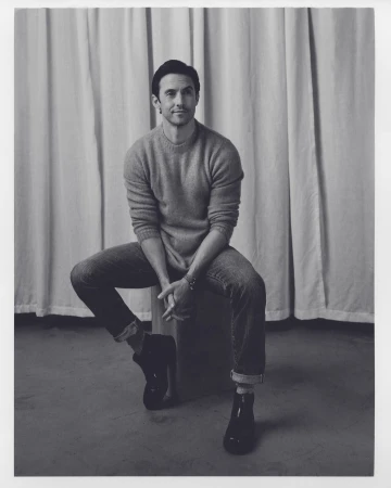 Portrait of a man in a crewneck sweater and cuffed jeans seated against a draped curtain background by Alex G. Harper