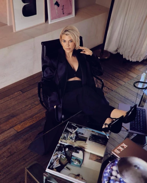 Stylish woman in a relaxed seated pose with a magazine on a glass table in a well-lit interior setting, wearing a black outfit.