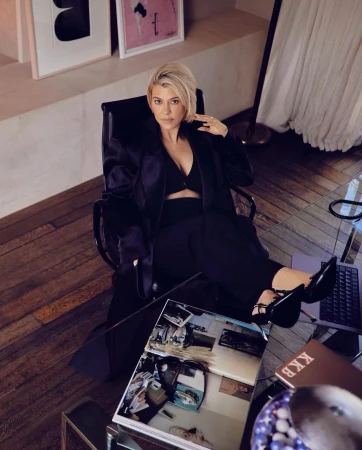 Stylish woman in a relaxed seated pose with a magazine on a glass table in a well-lit interior setting, wearing a black outfit.